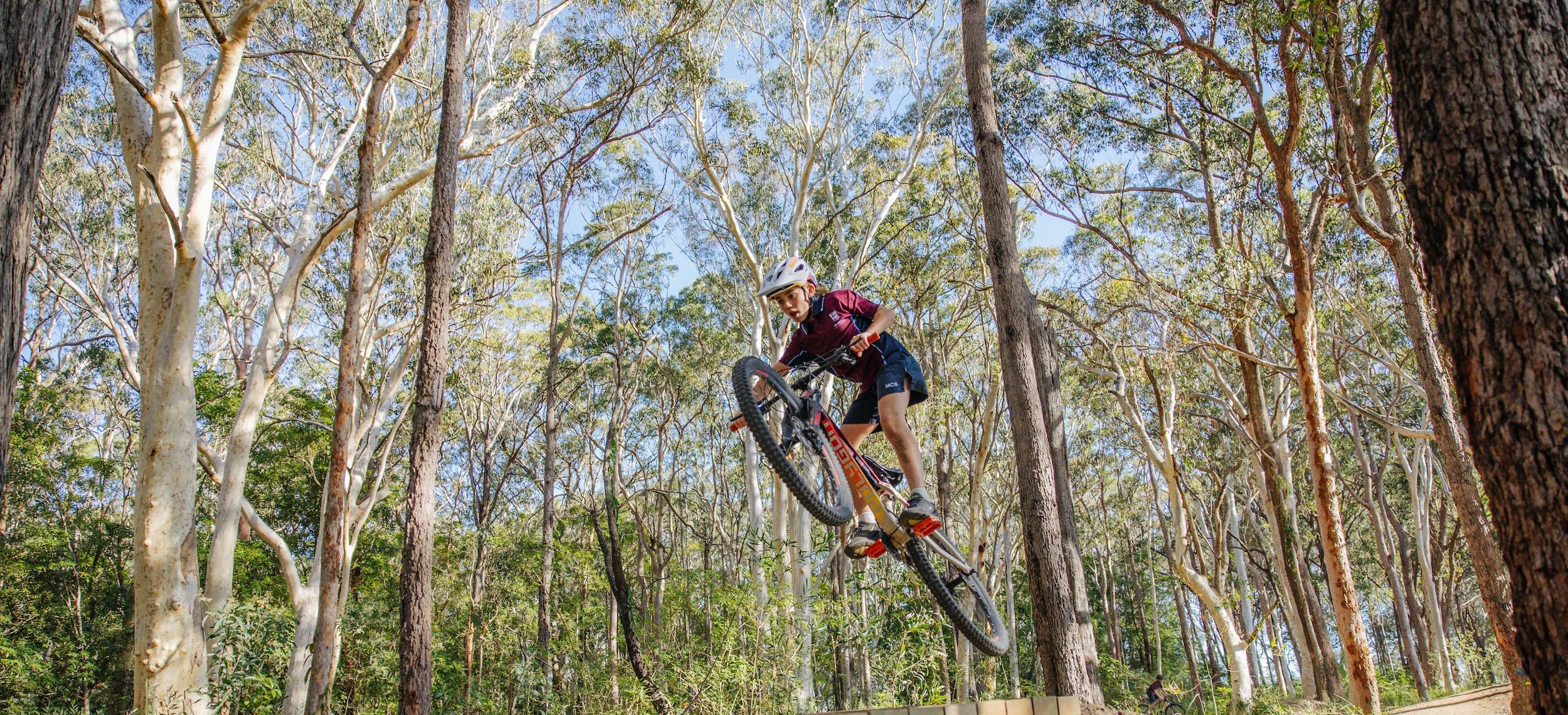 Low angle shot of female student jumping a mountain bike through a forest trail