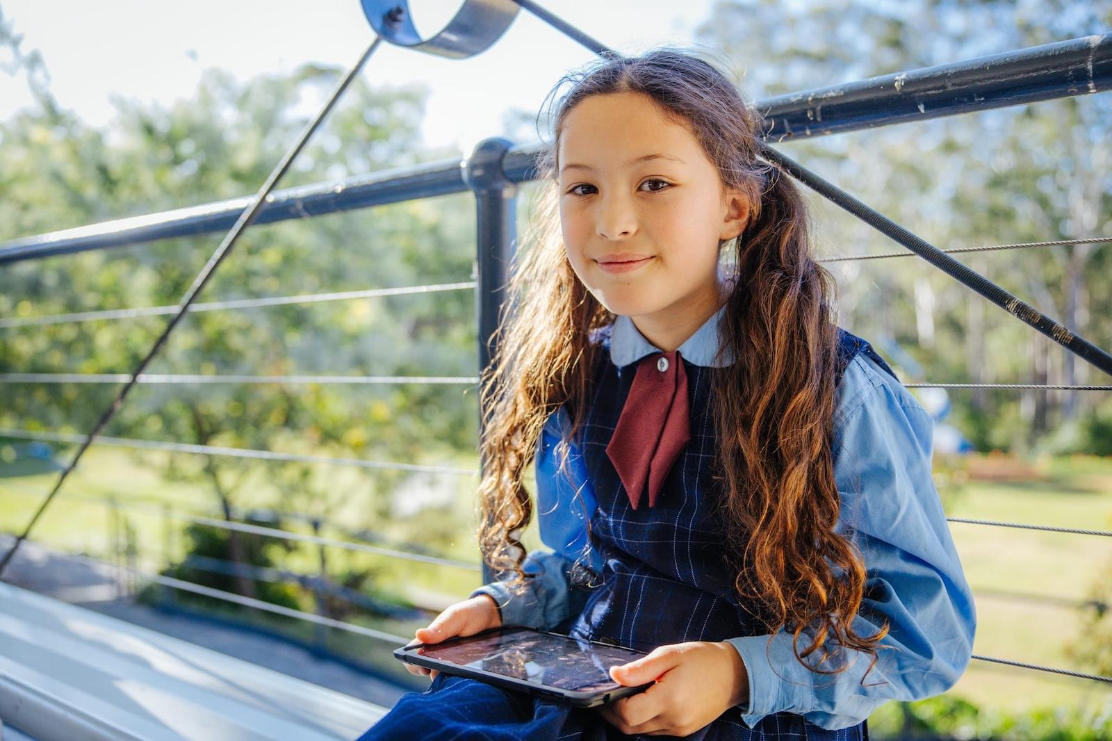 A student with her hair in pigtails sitting outdoors with a tablet, showcasing a supportive learning environment.